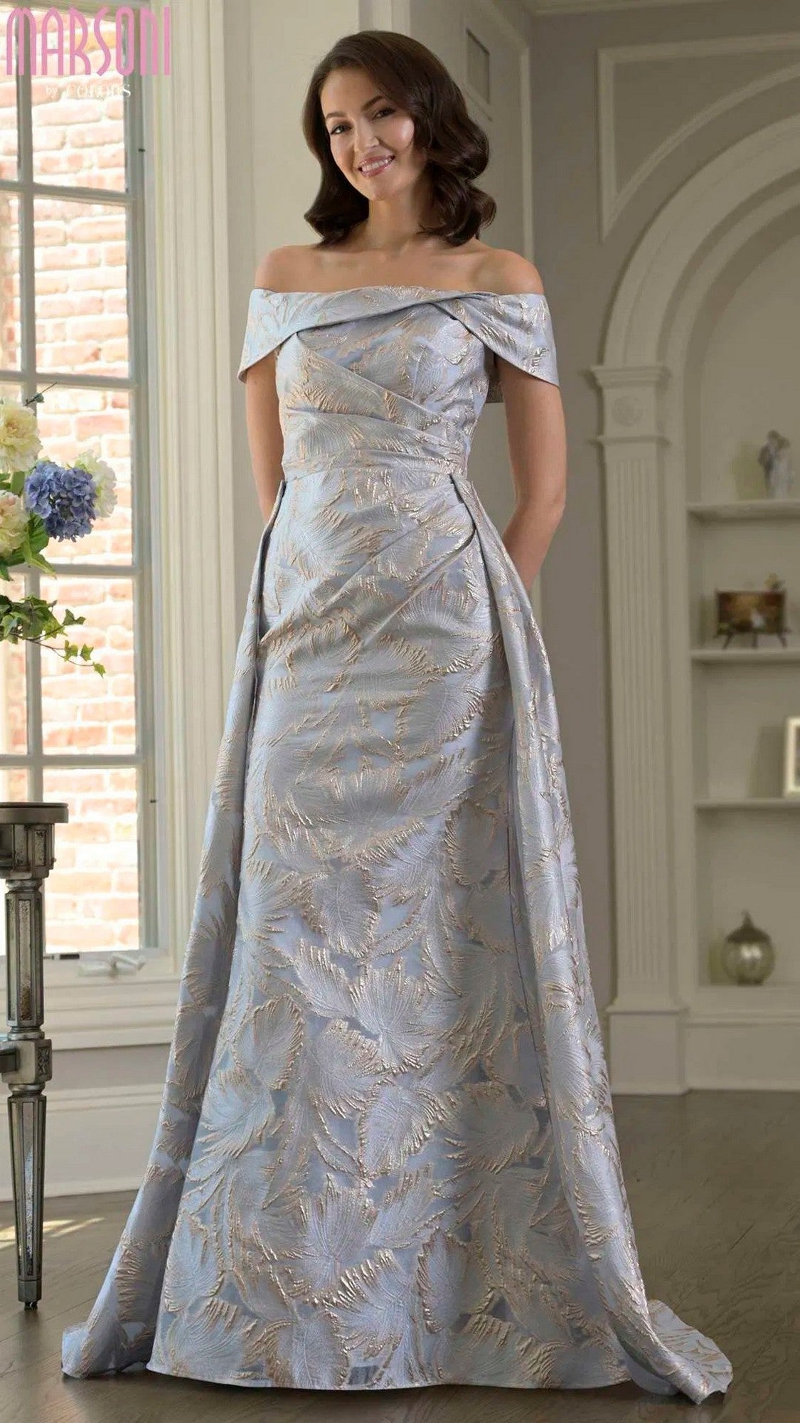 A woman stands indoors near a window and decorative shelves, wearing the Marsoni MV1382 long formal dress in Gold Blue—an off-the-shoulder, floor-length evening gown with a subtle leaf pattern.